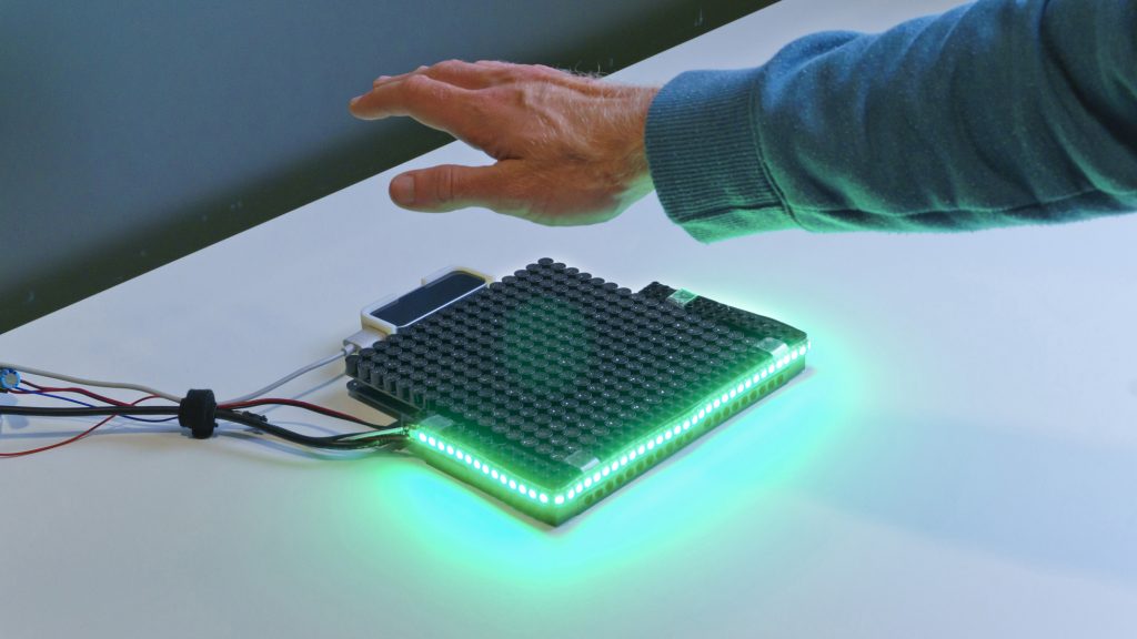 Photo of the HaptiGlow system. An Ultrahaptics UHEV1 device with a strip of LEDs around the front edge and left and right sides. The LEDs are green, indicating that the user has their hand in a good position.